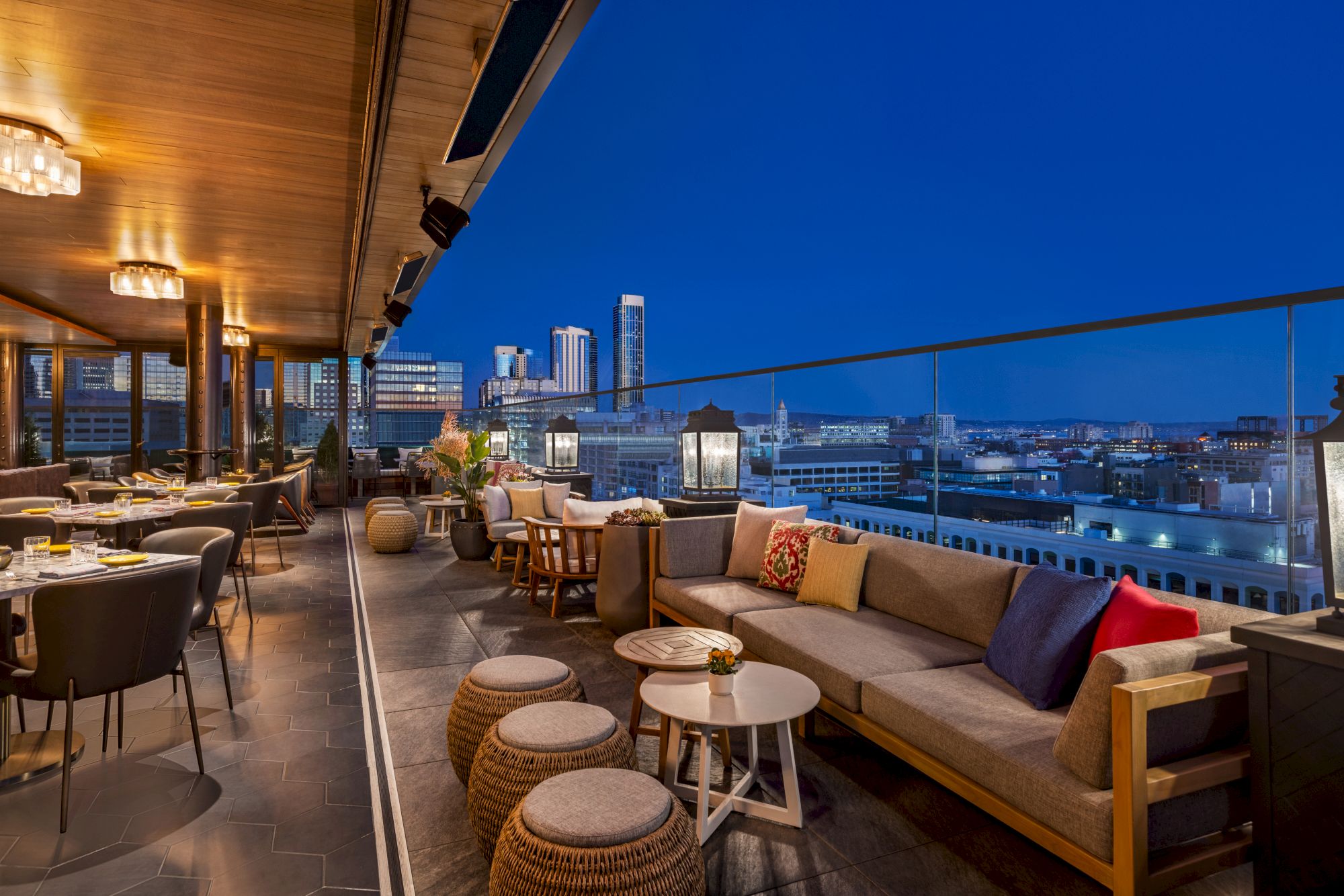 A rooftop terrace with a cozy seating area, tables, and city skyline views at dusk. The area is well-lit and has a modern, inviting ambiance.