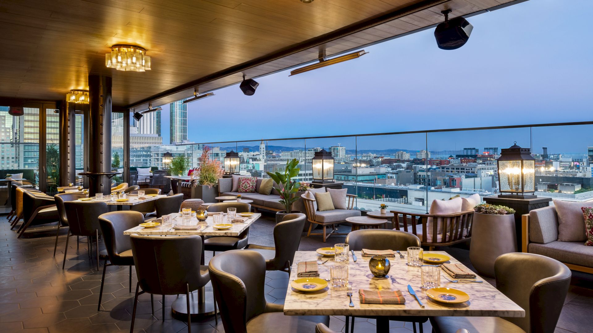 A rooftop restaurant with a city view, featuring elegant table settings, cushioned seating, and ambient lighting, ready for dining at dusk.