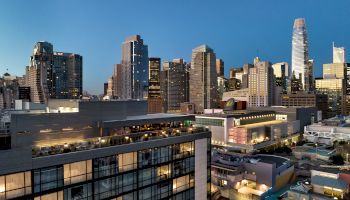 A city skyline at dusk with tall modern buildings, a mix of glass facades, and illuminated streets and rooftops in a bustling urban area.