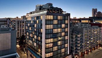 A modern glass-fronted building on a city street at dusk, with warm lights in the offices and surrounding mid-rise blocks.