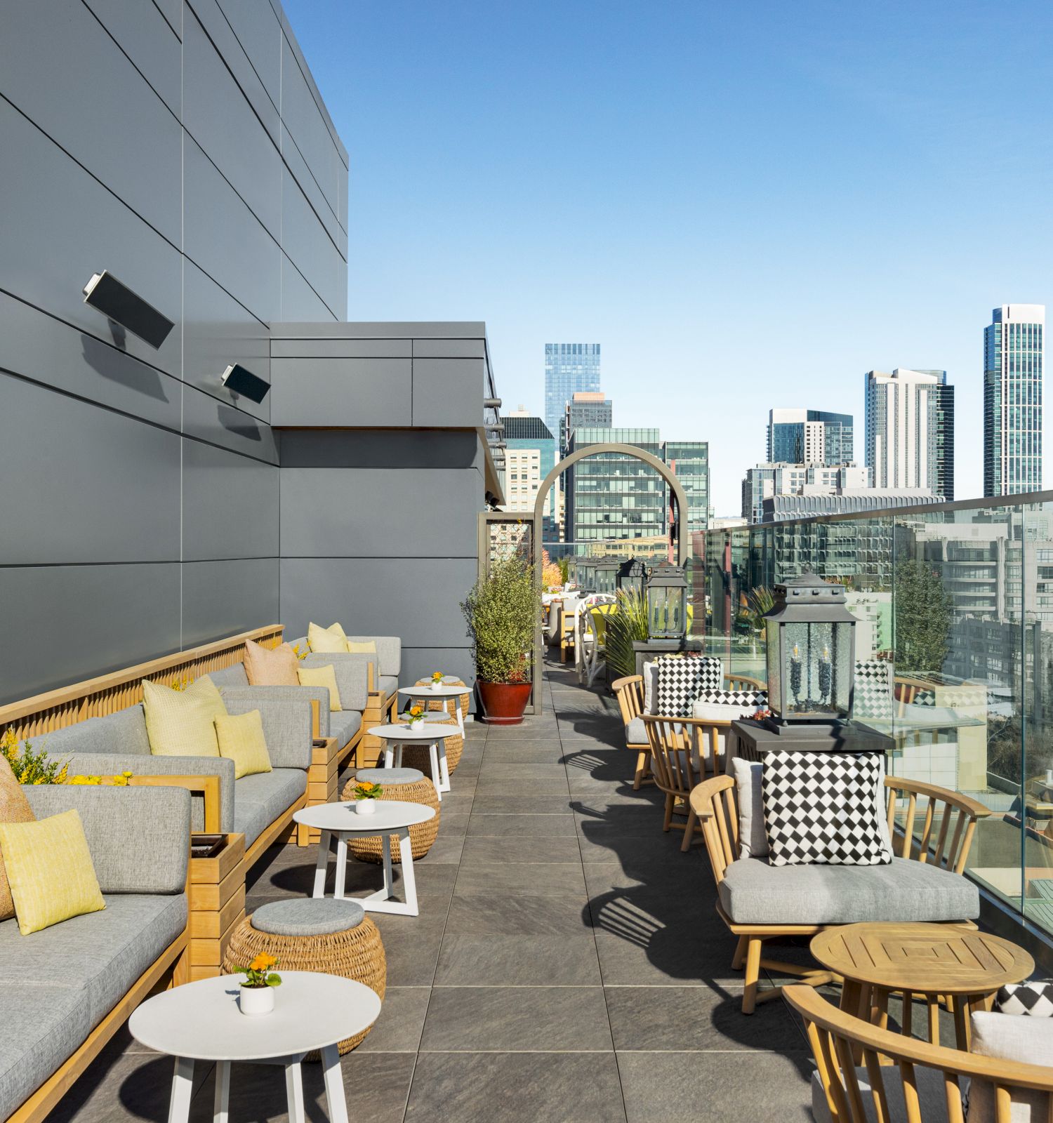 A modern rooftop terrace with comfortable seating, wooden furniture, and city views under a clear blue sky. Cityscape visible in the background.