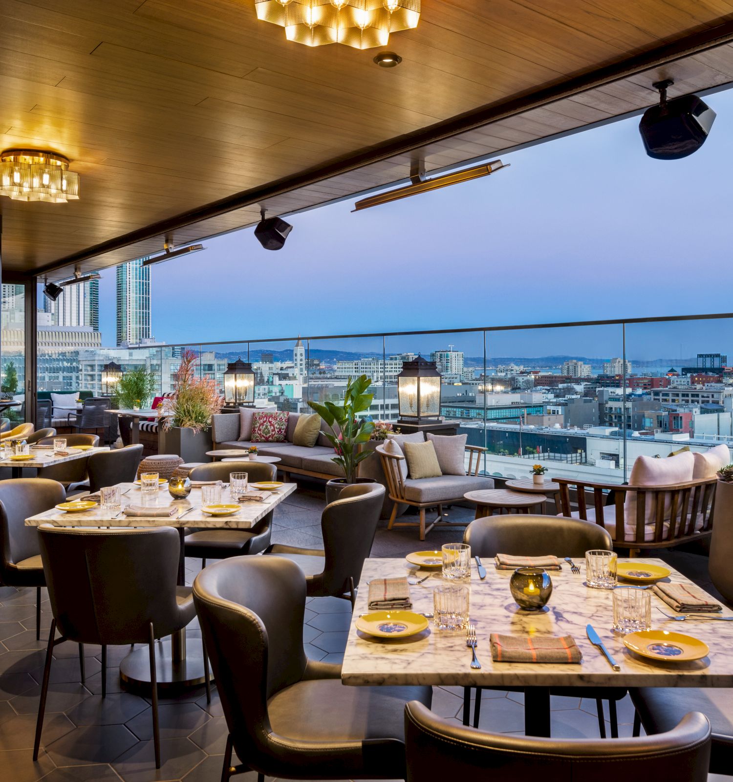 A stylish rooftop restaurant with well-set tables, elegant lighting, and a scenic view of a city skyline and waterfront in the background.