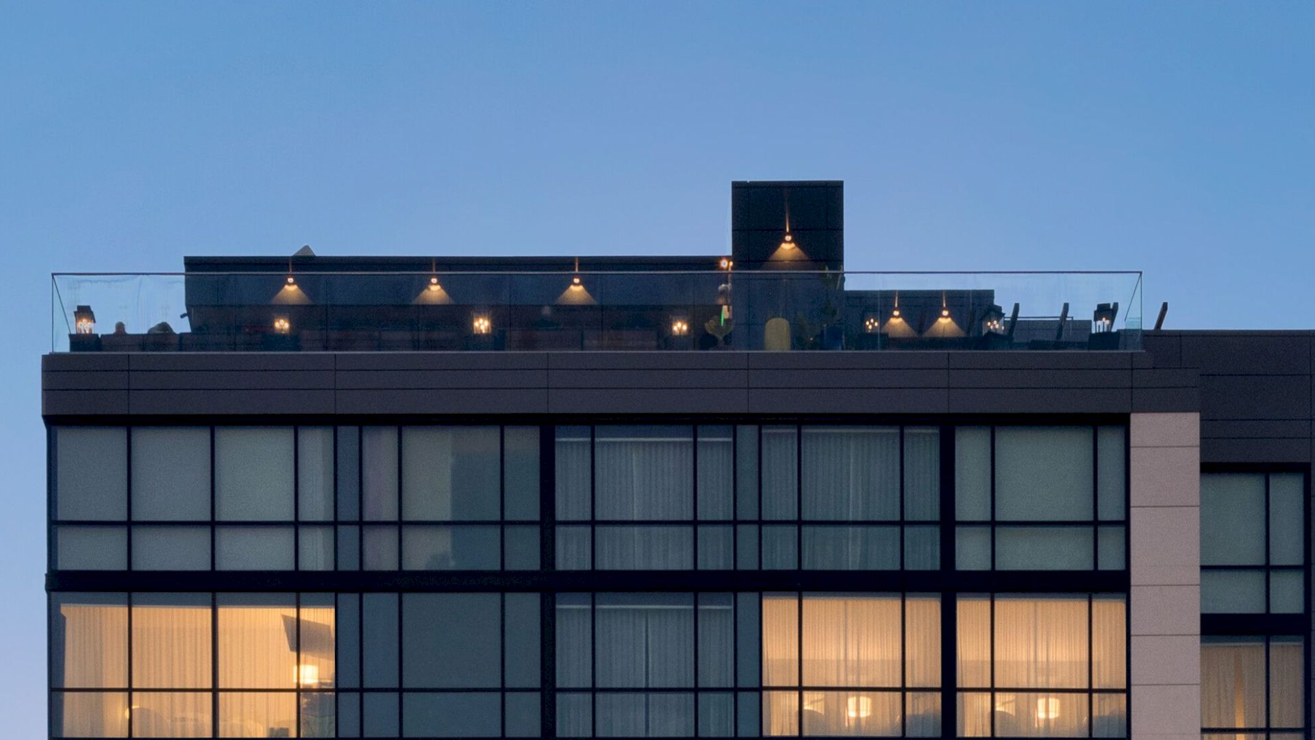 The image shows the top portion of a modern building with large windows and illuminated rooms at dusk, with a terrace on the rooftop.