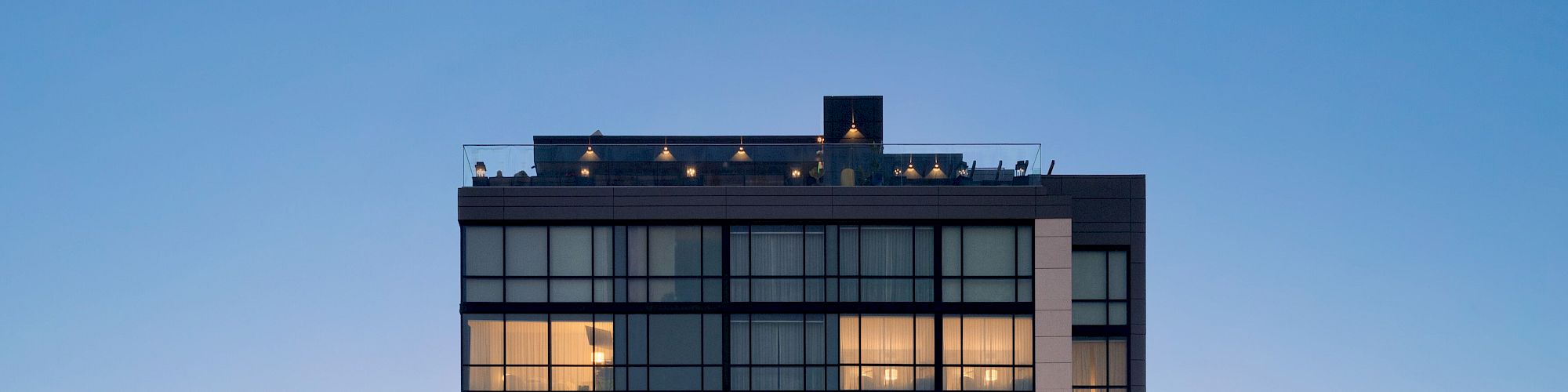 The image shows the top portion of a modern building with large windows and illuminated rooms at dusk, with a terrace on the rooftop.