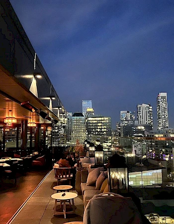 A rooftop bar with tables and chairs is overlooking a cityscape with illuminated skyscrapers under a twilight sky.