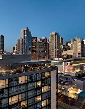 A city skyline at dusk with modern glass buildings, high-rises, and a busy urban area below, lights starting to glow as the evening sets in.