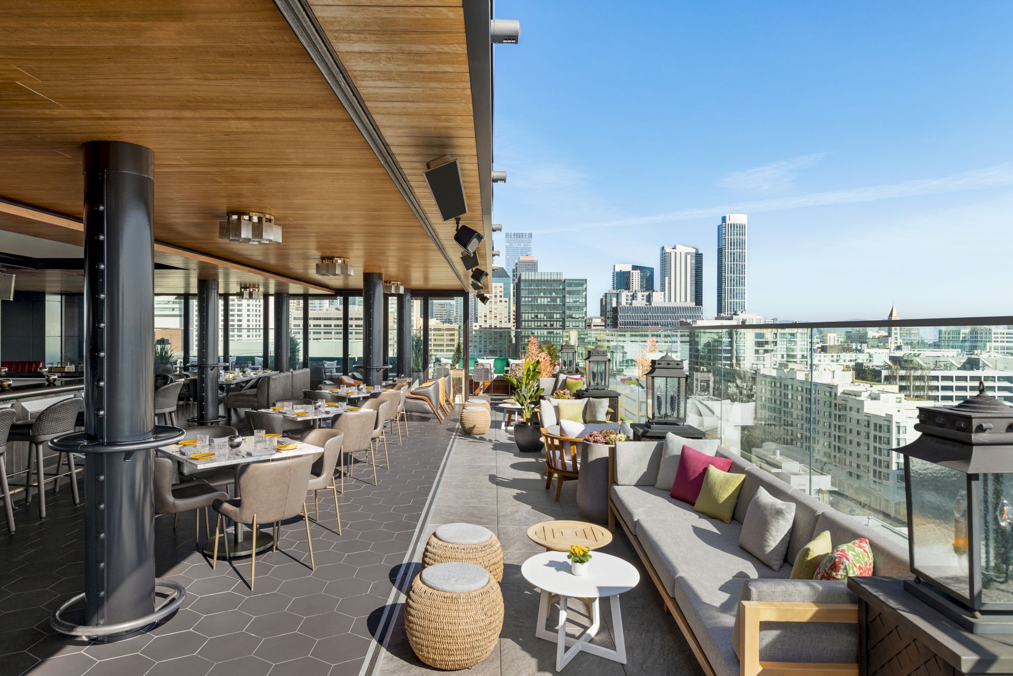 Rooftop lounge with modern seating, glass railing, city skyline, wicker chairs, round tables, and colorful cushions under a wooden canopy.
