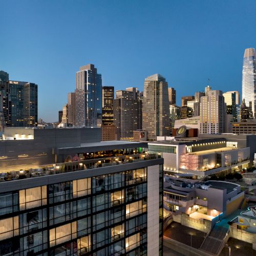 A city skyline at dusk with modern glass buildings, a rooftop terrace, and a gelid blue sky overhead.