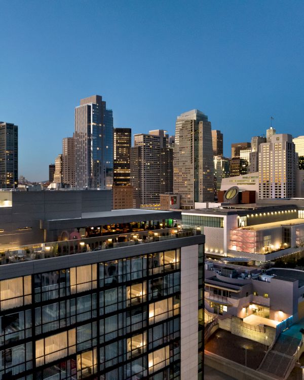 A city skyline at dusk with modern glass buildings, a rooftop terrace, and a gelid blue sky overhead.