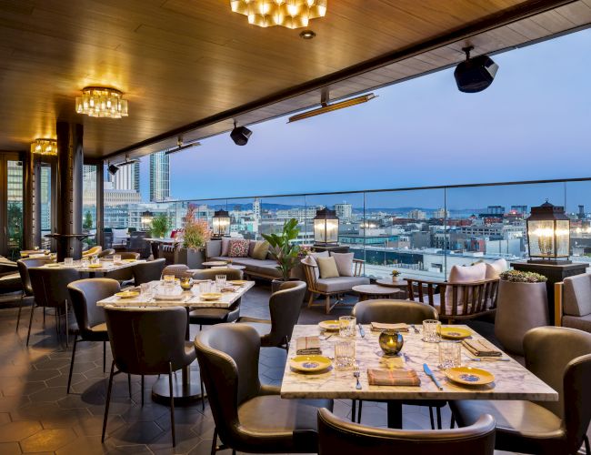 A stylish rooftop dining area with multiple tables set for dinner, warm lighting, and a cityscape view over the water at dusk.