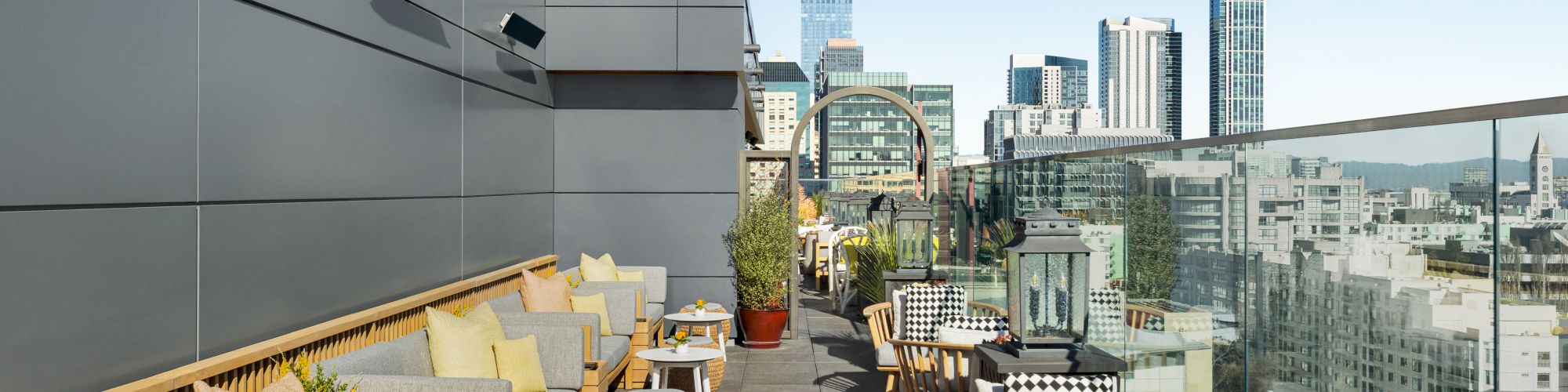 A stylish rooftop terrace with modern couches, small tables, and city viewsanta seating area along a glass railing.