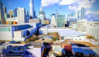 A bright aerial view of a dense city skyline with tall skyscrapers, mid-rise buildings, and a park-like complex under blue clouds.