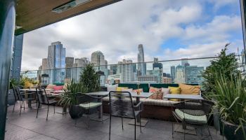 A modern rooftop patio with cushioned seating and city skyline views under cloudy skies, with plants lining the terrace, as sunlight fades.