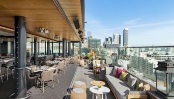 An outdoor rooftop dining area with modern seating, a glass railing, and views of the city skyline under a clear blue sky.