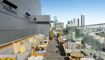 A modern rooftop terrace with outdoor seating, tables, and a glass railing, offering a city skyline view under a clear blue sky.