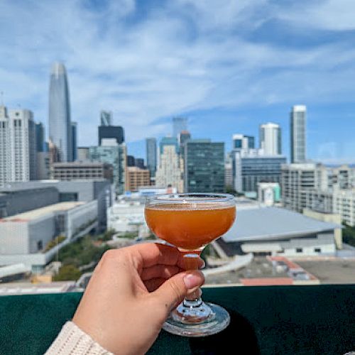 A person holds a small orange cocktail on a balcony with a city skyline and blue sky in the background.