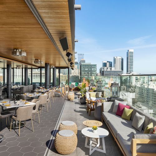 A stylish rooftop restaurant with seating, lounge sofas, and small tables, overlooking a city skyline with modern glass buildings.