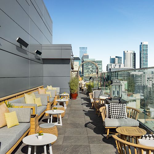 A rooftop terrace with loungers, tables, and potted plants overlooking a city skyline, sunny and modern.