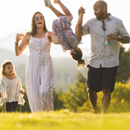 A family of four walking outside; the parents hold the hands of a girl who's mid-air, while another child walks beside them, all look happy.