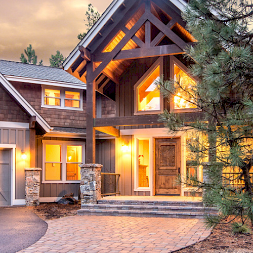 A large, beautifully lit modern house with a stone pathway and garage, surrounded by trees during dusk.