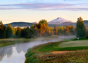 The image shows a serene golf course beside a river with mist rising, surrounded by trees and mountains, with a snow-capped peak in the background.