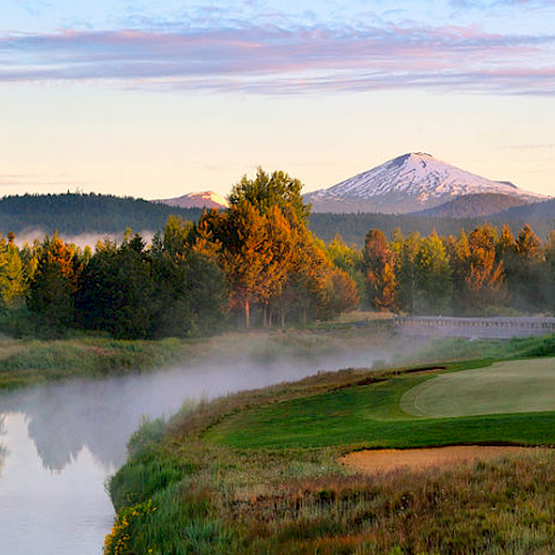 The image shows a serene golf course beside a river with mist rising, surrounded by trees and mountains, with a snow-capped peak in the background.