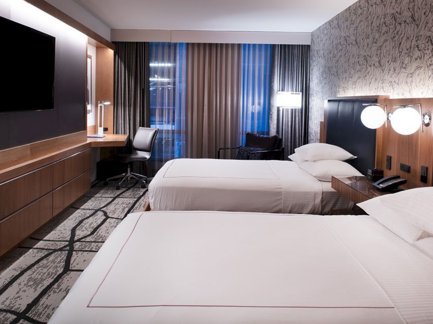 The image shows a modern hotel room with two double beds, a TV, a desk with a chair, and stylish decor.