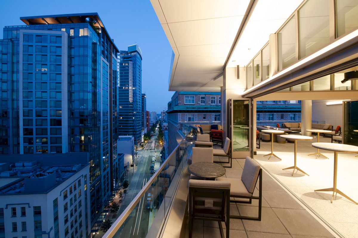 A modern balcony overlooking a cityscape at dusk, with tables and chairs arranged for seating and a view of tall buildings.