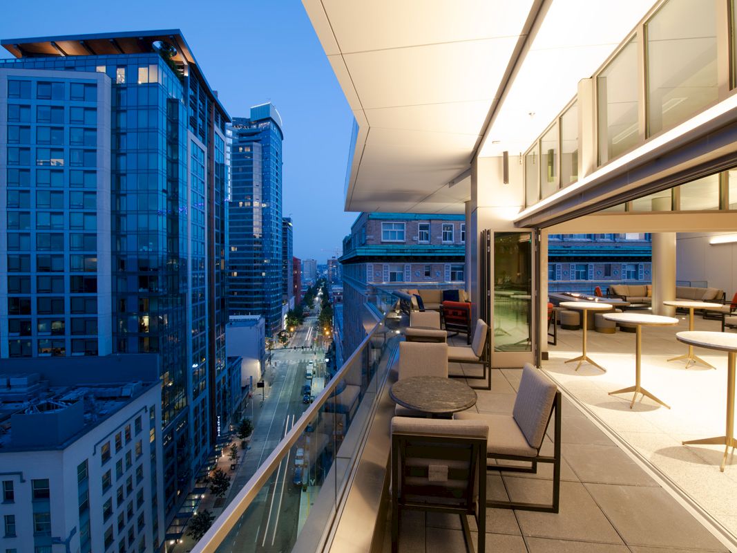 A modern balcony overlooking a cityscape at dusk, with tables and chairs arranged for seating and a view of tall buildings.