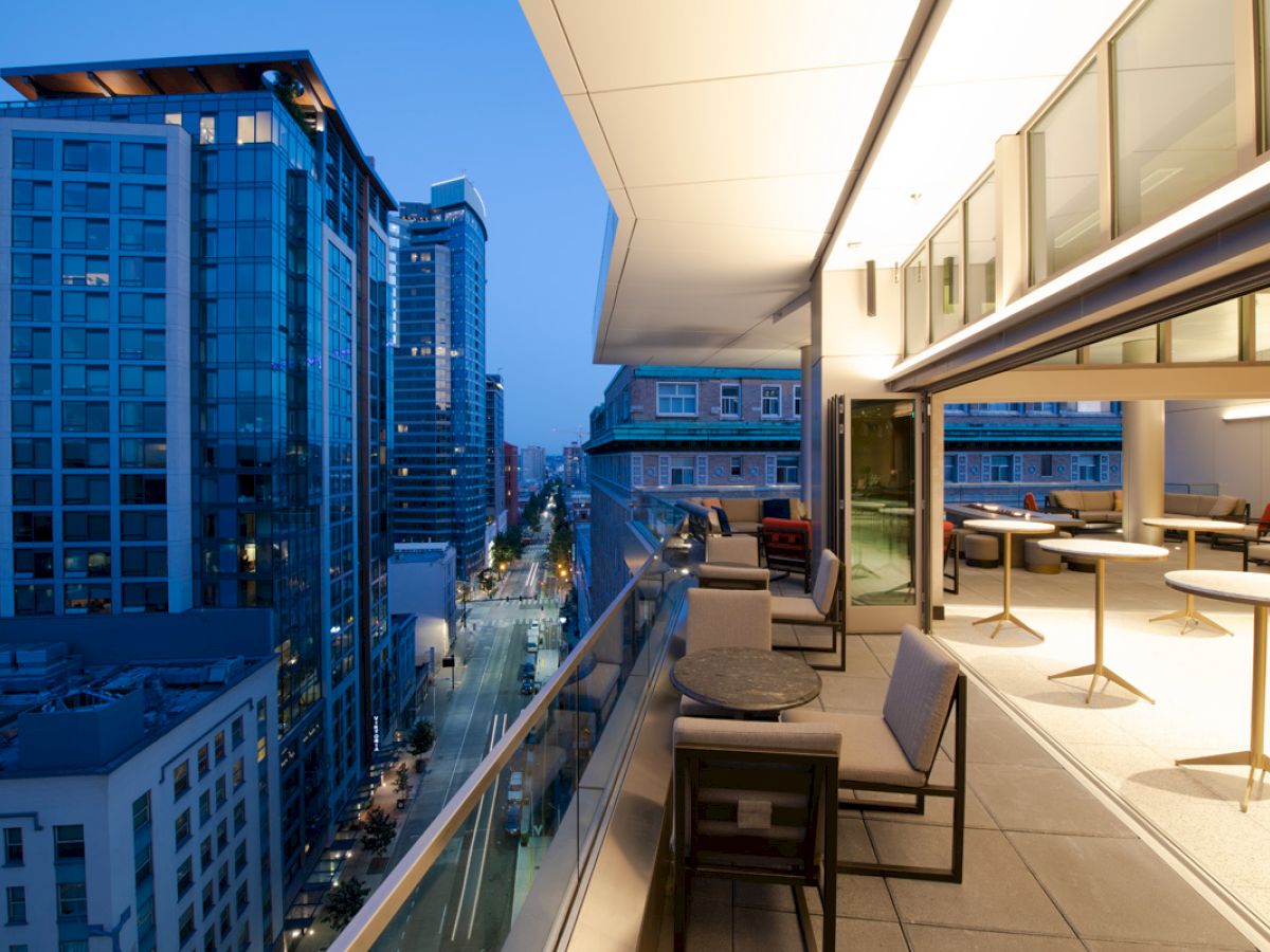 The image shows a modern balcony or terrace at dusk, overlooking a cityscape with tall buildings and a street below.