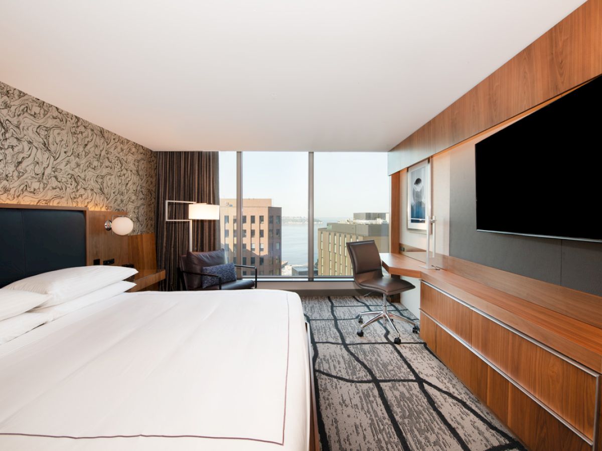 The image shows a modern hotel room with a large bed, a desk, a chair, a flatscreen TV, and a city view through a wide window.
