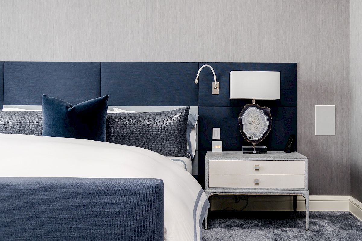 A modern bedroom with a navy and white color scheme, featuring a bed, navy headboard, pillows, bedside table, lamp, and wall-mounted reading light.