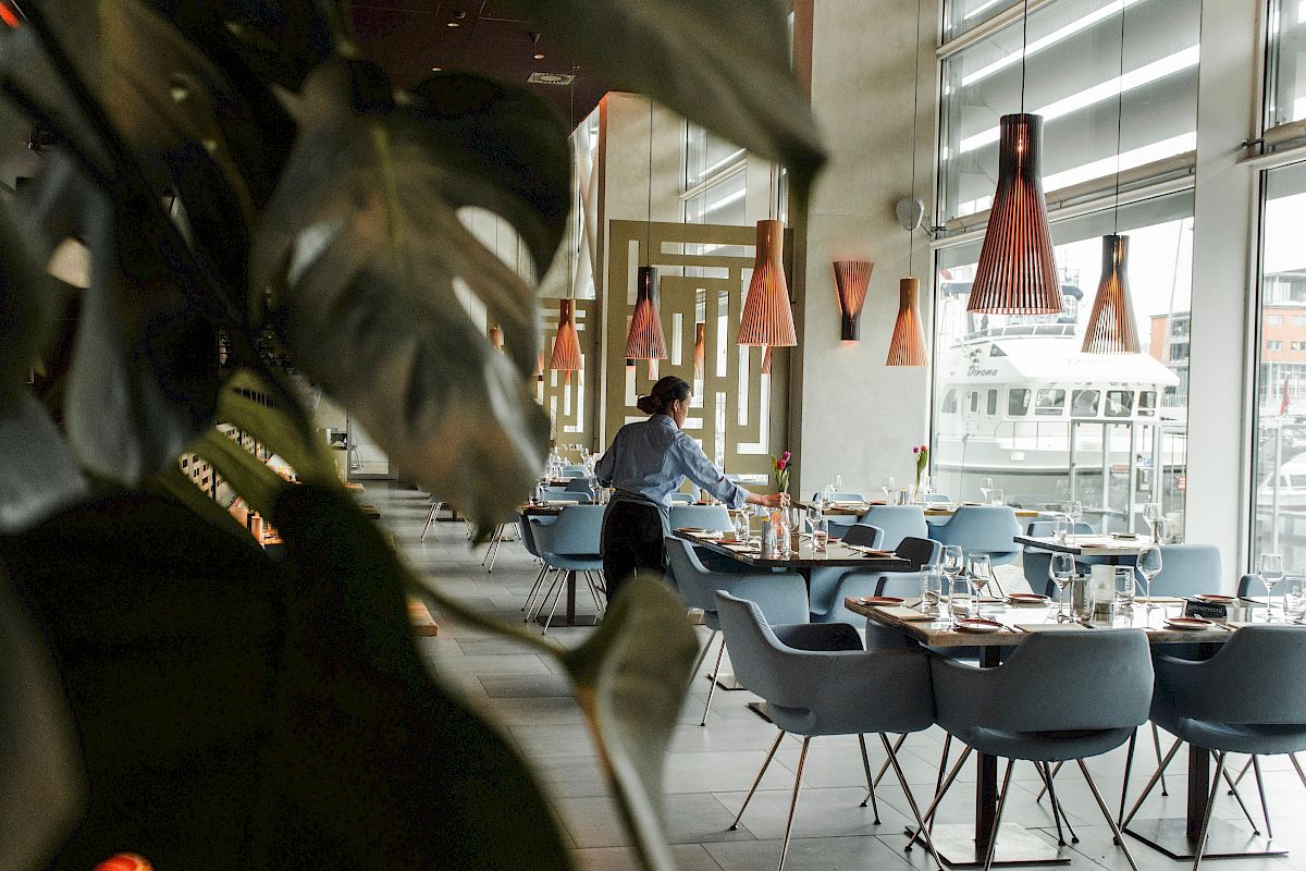 A person is setting up tables in a modern restaurant with stylish lighting and large windows, viewed through plant leaves in the foreground.