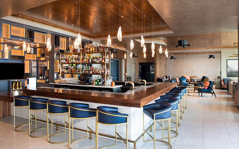 A stylish bar with a wooden counter, blue stools, and a well-stocked liquor shelf, featuring modern lighting and a cozy seating area in the background.