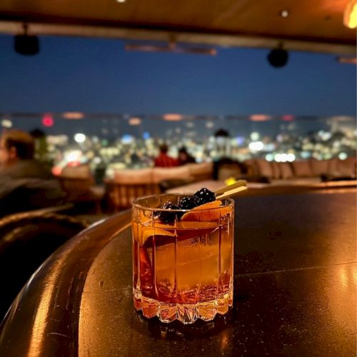 A cocktail is on a bar counter with a scenic city view in the background at night, featuring warm ambient lighting and an elegant atmosphere.