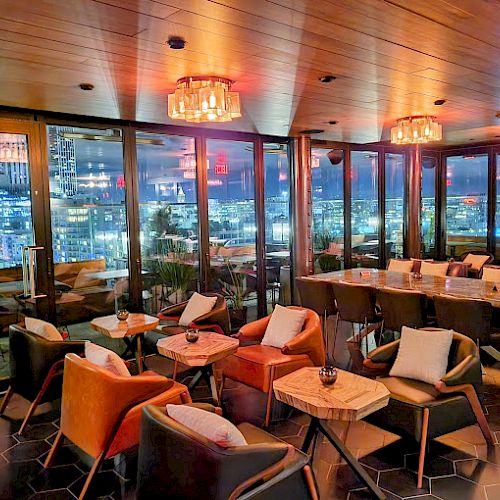 A modern rooftop lounge with cozy seating arrangements, illuminated lighting, large windows showcasing a cityscape view, and tables for dining.