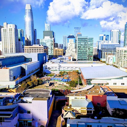 Image of a city skyline with modern skyscrapers and various buildings under a partly cloudy sky, viewed from an elevated position.