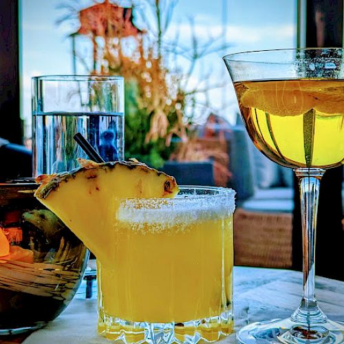 The image shows three drinks on a table: a cocktail with a lemon slice, a pineapple-decorated yellow drink, and a glass of clear water.