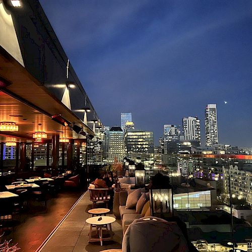 A rooftop bar with seating areas overlooks a modern city skyline at night, featuring lit buildings and a clear sky.