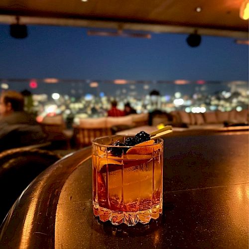 A close-up of a cocktail on a bar counter, with an evening cityscape and blurred skyline visible in the background, in a dimly lit setting.