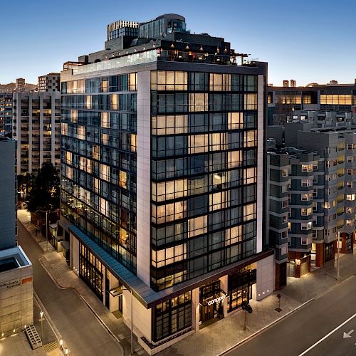 A tall, modern building with many windows stands illuminated in an urban area at dusk, surrounded by other buildings and a street lined with trees.