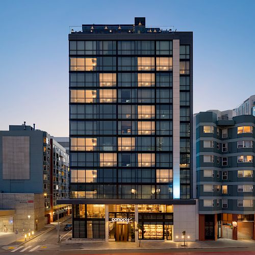 The image depicts a modern, multi-story building with illuminated windows situated between other buildings in an urban setting during dusk.