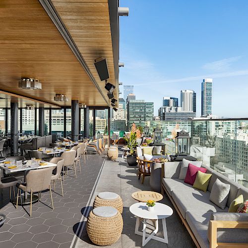 An outdoor rooftop terrace with seating areas, tables, and city views. The space has modern decor, including cushions and stools, under a wooden canopy.